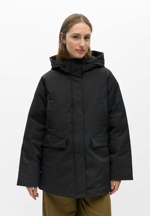 Femme portant une veste d'hiver noire avec capuche et poches avant, se tenant devant un fond blanc uni.