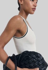 Schwarze geflochtene Lederhandtasche mit doppelten Griffen, gehalten neben einem hellen, gerippten Tanktop mit schwarzem Rand an den Trägern und dem Ausschnitt.