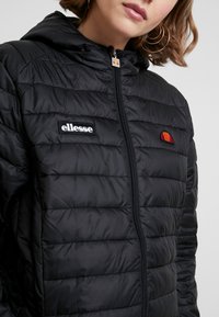 Veste rembourrée noire avec capuche, dotée de coutures horizontales, fermeture éclair et logos brodés sur la poitrine. Texture lisse.