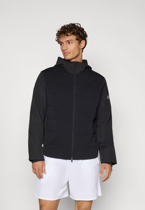 Veste noire à capuche avec un torse rembourré, des manches lisses, une fermeture éclair et un logo discret sur le bras gauche. Tissu doux et texturé.