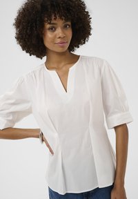 Bluză albă, cu mâneci scurte și decolteu în V, cu detalii plisate la mâneci. Confecționată dintr-un material neted, cu plisse pe laterale, purtată peste blugi.