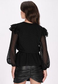 Zwarte blouse met stippenpatroon, doorzichtige lange mouwen met rimpel- schouders, gecombineerd met een zwarte leren rok met geplooide details.