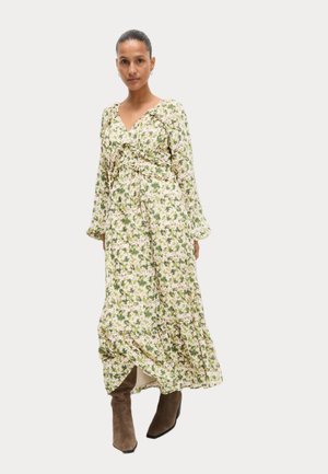 Robe maxi fleurie à manches longues, fond crème, motif floral vert et marron, taille cintrée et ourlet froncé. Bottes marron jusqu'aux genoux.