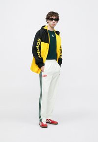 Κίτρινο και μαύρο αδιάβροχο με το λογότυπο "adidas", πράσινο μπλουζάκι, άσπρο παντελόνι με πράσινες ρίγες, κόκκινα παπούτσια και σκούρα γυαλιά ηλίου.