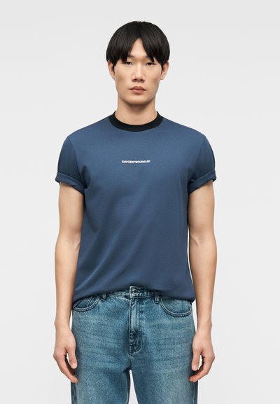 Άνδρας που στέκεται προς τα εμπρός φορώντας μπλε μπλουζάκι Emporio Armani με μανίκια γυρισμένα και ανοιχτόχρωμο μπλε τζιν μπροστά σε απλό φόντο.