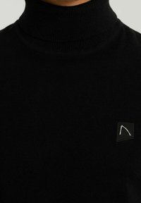 Schwarzer Rollkragenpullover aus weichem Stoff, mit geripptem Kragen und einem kleinen Logopatch in der oberen linken Ecke.