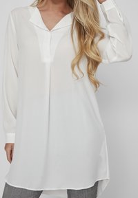 Vit långärmad blus med V-ringning, tillverkad av ett mjukt och lätt material och med en något böjd fåll, matchad med rutiga byxor.