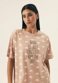 Jasnoróżowa koszulka z krótkim rękawem, w białe wzory chmur, z centralnym nadrukiem tekstowym "DESZCZ TO TYLKO KONTETY Z NIEBA."
