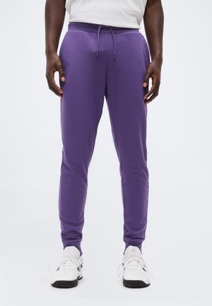 Mężczyzna w fioletowych spodniach dresowych ze sznurkiem w talii, białych trampkach i białej koszuli, stojący na jednolitym jasnym tle.