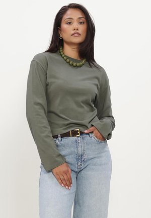 Kvinde iført olivengrøn langærmet top, blå jeans med brun bælte, grøn perlehalskæde og gyldne creoløreringe, poserer med den ene hånd i lommen.