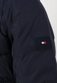 Granatowa kurtka puchowa z pikowanym wzorem, z czarną naszywką z napisem "TOMMY HILFIGER" oraz poziomym akcentem w kolorach czerwonym, białym i niebieskim.