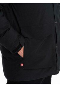 Persona che indossa una giacca nera con una grande tasca frontale, mano all'interno della tasca e cinghia in Velcro sul polsino della manica.