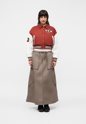 Frau trägt eine rot-weiße Collegejacke mit der Nummer 3 auf dem Ärmel, einen langen beigen Rock mit großen Taschen und schwarze Schuhe und steht vor weißem Hintergrund.