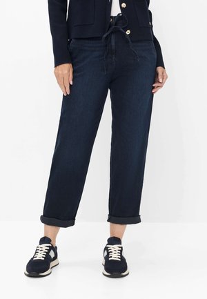 Person iført opkørte mørkeblå jeans, navyblå jakke med guldknapper og navyblå sneakers med hvide snørebånd og såler.