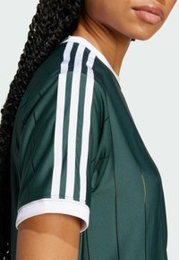 Zielony sportowy trykot z białymi paskami na ramionach, krótkimi rękawkami i prążkowanym kołnierzykiem. Gładka faktura z pionowymi przeszyciami.