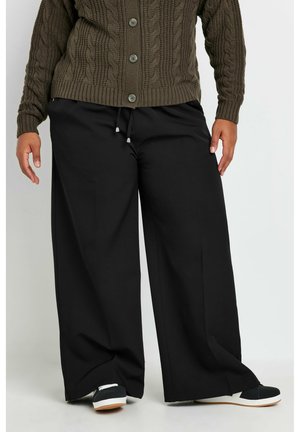 Zwarte wijde broek met een elastische koordjes taille en twee zijzakken, gecombineerd met een gestructureerde olijfgroene kabelgebreide cardigan en sneakers.