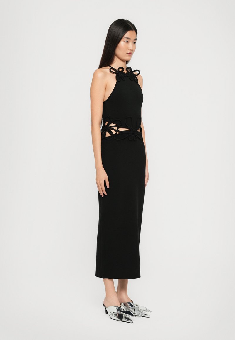 Robe noire en halter avec des découpes et des boucles décoratives à la taille. Associée à des mules argentées à bout pointu et à talon bas.