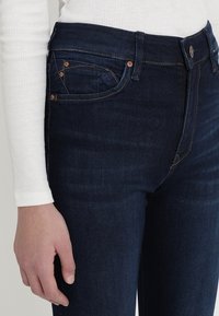 Jeans de mezclilla azul oscuro con un diseño ajustado, cintura alta, bolsillos frontales prominentes y acentos de hardware cobre en las costuras y botones.