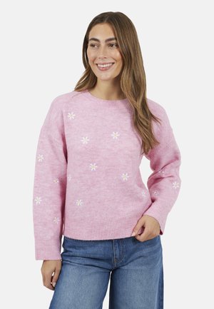 Donna sorridente con capelli lunghi che indossa un maglione rosa ricamato con margherite bianche e jeans blu, posando con una mano sui fianchi.