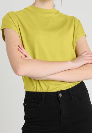 Persoon draagt een geel T-shirt met korte mouwen en zwarte broek, met gekruiste armen over de borst tegen een effen achtergrond.