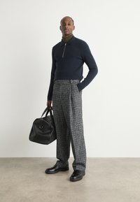 Pulover tricotat bleumarin cu guler cu fermoar, pantaloni cadrila gri cu o croială lejeră, geantă din piele neagră și pantofi închiși la culoare. Textură netedă, design modern.