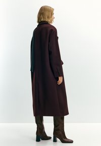 Vrouw staand in profiel met een lange donker bordeauxrode jas, bruine laarzen met hak en een sjaal over haar schouder gedrapeerd tegen een eenvoudige achtergrond.