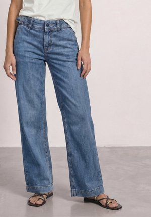 Person iført løse blå jeans, hvid top og sorte åbne sandaler, der står på en glat gulv mod en ensfarvet lys baggrund.