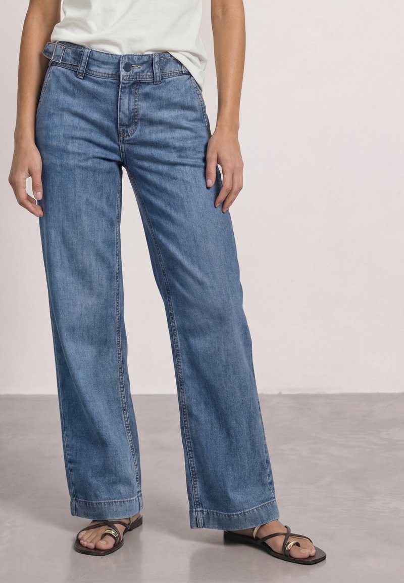 Person wearing loose blue jeans, white top, and black open-toe sandals standing on a smooth floor against a plain light background.