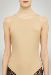 Costume da bagno senza maniche di colore nude con un corpetto liscio e sagomato, arricchito da dettagli in pizzo nella parte inferiore. Presenta una scollatura rotonda.