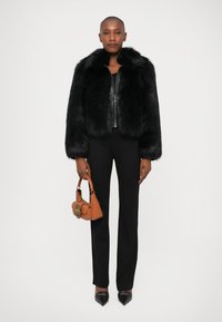 Schwarze Kunstfelljacke mit einem Kragen, getragen über einem schwarzen Oberteil und Hosen. Das Model hält eine kleine orange Handtasche mit Metallbeschlägen. Spitz zulaufende schwarze Schuhe.