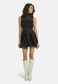 Robe mini noire avec un corsage ajusté, un décolleté transparent et une jupe volumineuse. Associée à de hautes bottes blanches à talons avec un motif crocodile.