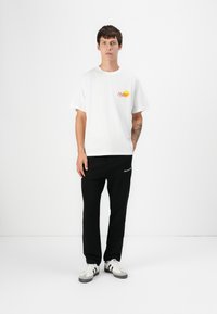 T-shirt bianca in cotone con grafica rossa "Holiday", abbinata a pantaloni sportivi neri con dettagli ricamati, completata da sneakers bianche e nere.