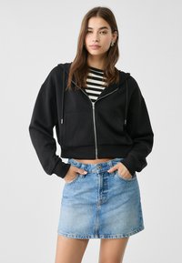 Felpa nera cropped con zip frontale e due tasche, abbinata a una mini gonna in denim azzurro. Camicia a righe a maniche lunghe sotto.