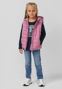 Roze puffer vest met een capuchon over een donkerblauw overhemd met lange mouwen met een bloemmotief, gecombineerd met blauwe jeans en witte sneakers.