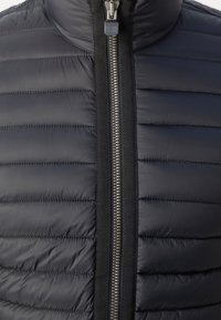 Gros plan d'une veste matelassée noire avec des coutures horizontales et une fermeture éclair métallique centrale partiellement fermée.