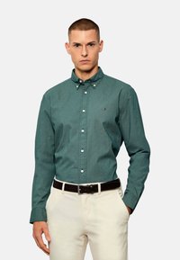 Camisa de botones verde con estampado, cuello puntiagudo, botones blancos y un logo en el pecho, combinada con pantalones de color claro y un cinturón negro.