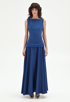 Donna con capelli ricci che indossa un top senza maniche di colore blu reale e una gonna lunga abbinata, in piedi davanti a uno sfondo bianco.