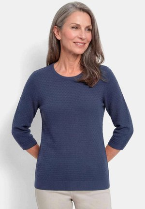 Frau mit grauen Haaren lächelt, trägt einen strukturierten marineblauen Pullover mit Dreiviertelärmeln und helle Hosen vor einfachem Hintergrund.
