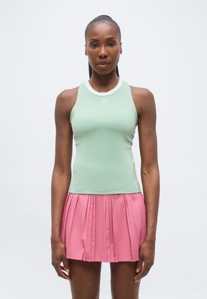 Kvinde stående med front mod kamera iført en ærmeløs lysegrøn top og en lyserød plisseret nederdel mod en ensfarvet baggrund.