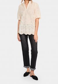 Blouse crème en broderie anglaise avec des manches courtes, un ourlet festonné et une légère silhouette peplum, associée à un jean noir retroussé et des chaussures plates noires.