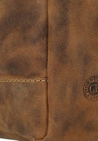 Brown leather bag featuring a textured surface with a visible seam and a circular logo embossed on the side. Simple design and sturdy construction.