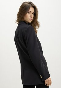 Schwarzer maßgeschneiderter Blazer mit einem leicht strukturierten Design, Reverskragen und metallischen Knöpfen an den Ärmeln. Glattes Material und taillierte Form.