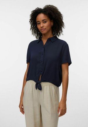 Kvinde med krøllet hår iført en navy kortærmet skjorte med knapper, bundet i taljen, og beige løstsiddende bukser, stående foran en hvid baggrund.