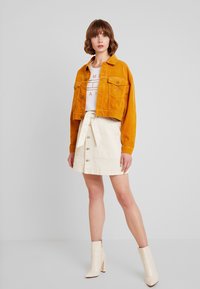 Senapsgul kordfløyelsjacka med två bröstfickor, vit t-shirt med grafik, beige knäppkjol och krämfärgade klackstövletter.