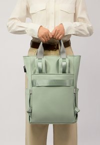 Borsa tote verde menta con doppio manico superiore, tessuto strutturato e tasche laterali. Presenta una cinghia piatta sulla parte anteriore per un tocco di dettaglio in più.