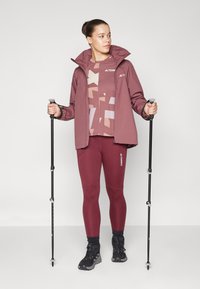 Chaqueta impermeable color burdeos con capucha, blusa estampada y leggings a juego. Botas de senderismo negras y bastones de trekking complementan el atuendo.