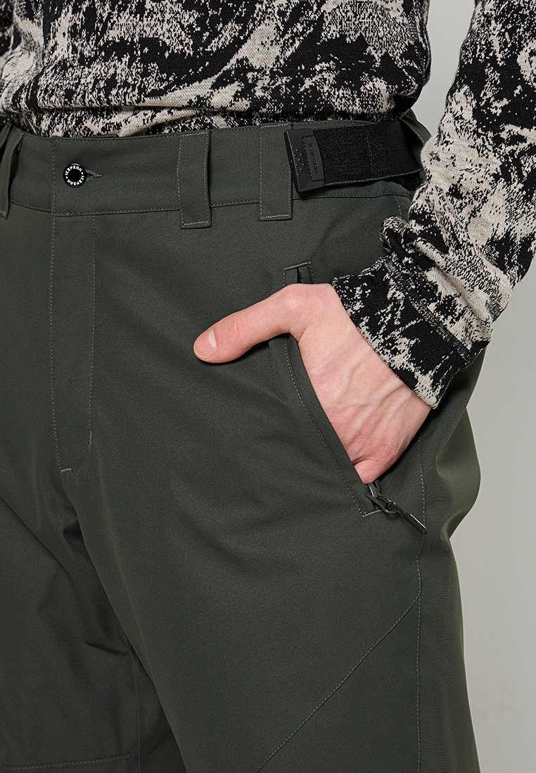 Grüne, weiche Shell-Hosen mit schmaler Passform, mehreren Taschen und einem strukturierten Bund sowie einem akzentuierten Reißverschlussdetail auf der Hüfte.