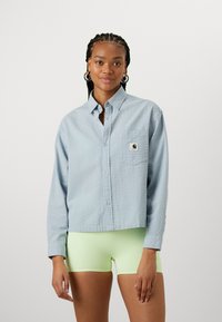 Ljusblå, långärmad skjorta med knappar med ett texturerat mönster och en liten bröstficka med en logotyp. Bärs över neongröna shorts.
