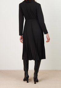 Černé plisované šaty s dlouhými rukávy a páskem v pase. Textura látky je hladká. Nositelná s černými kozačkami na podpatku po kolena.