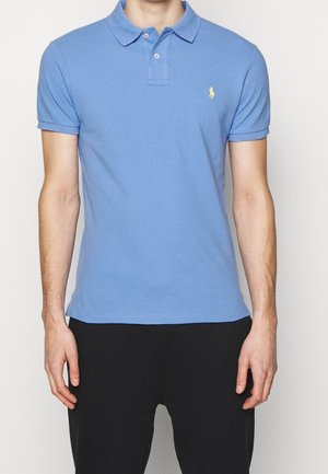 Man wearing a light blue short-sleeve polo shirt with a small yellow logo on the chest and black pants, standing against a white background.